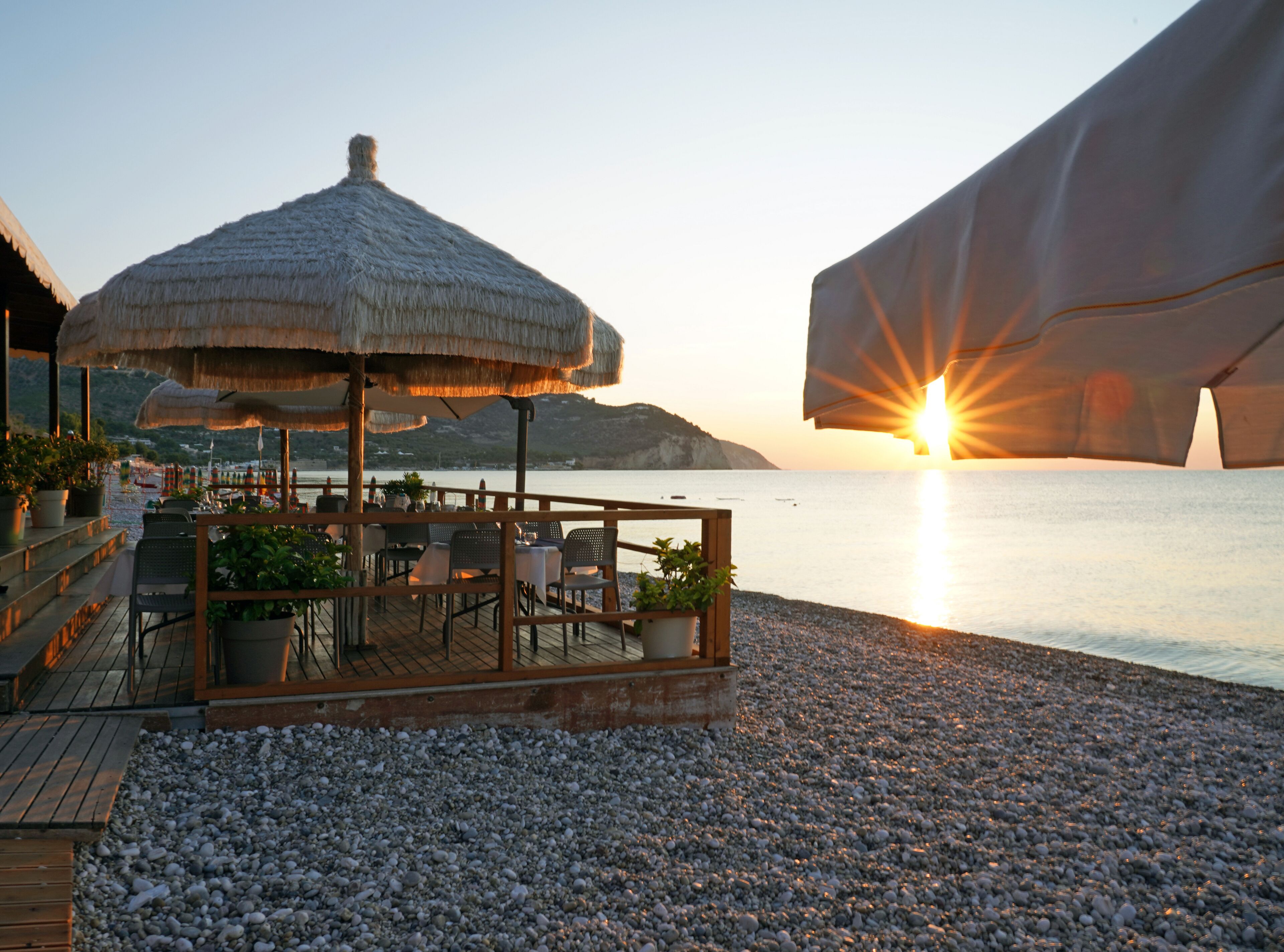 Un restaurant en bord de mer, doté d’un parasol en chaume, d’une terrasse en bois et de plantes en pot.