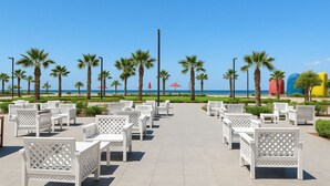 Une terrasse au bord de la plage, avec des meubles d’extérieur blancs, des palmiers et une vue dégagée sur l’océan.