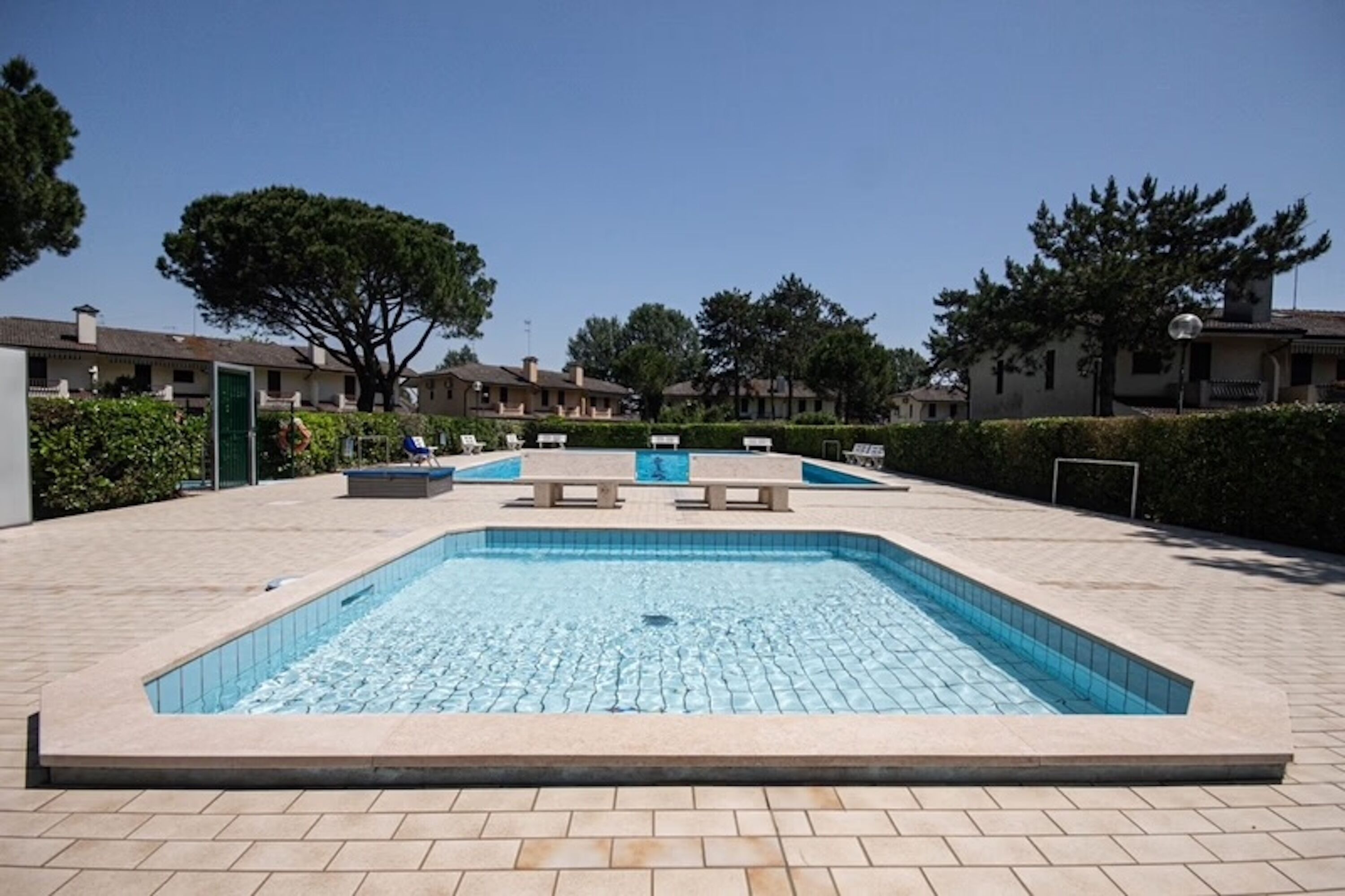 A rectangular swimming pool with a tiled deck, surrounded by benches and a hedge.