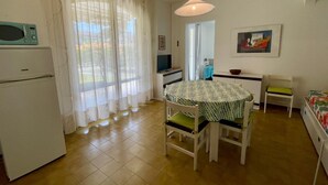A compact kitchen with a refrigerator, a dining table with chairs, a hanging light fixture, and a painting on the wall. - Holiday Home With Garden and Shared Pool (Caorle)