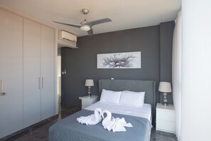 A hotel room with a bed, two bedside tables with lamps, a ceiling fan, an air conditioning unit, a gray wall, and a large floral artwork. - Protaras Holiday Villa Bp6 (Paralimni)