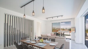 A modern dining area with a wooden table set for six, surrounded by chairs. There is a sofa and a dining table with a city view through large windows. - Protaras Vie Bleu Villa Vb4 (Paralimni)