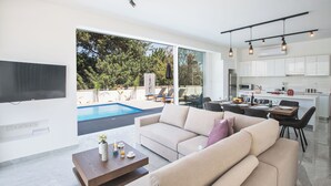 A modern living room with a large sofa, a coffee table, and a TV. There is a dining area with a table and chairs, and a kitchen with cabinets and appliances. A pool is visible through the glass doors. - Protaras Vie Bleu Villa Vb4 (Paralimni)
