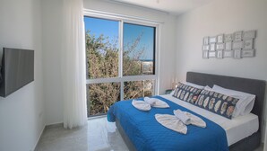 A bedroom with a large window, a bed with blue bedding, and a wall-mounted TV. - Protaras Vie Bleu Villa Vb4 (Paralimni)