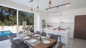 A modern kitchen with white cabinets, a dining area with a table set for four, and a pool visible through the glass doors. - Protaras Vie Bleu Villa Vb4 (Paralimni)