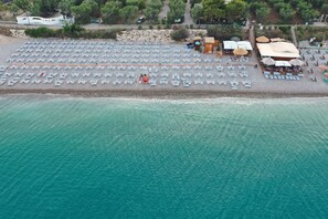 An aerial view of a beach with rows of sun loungers and umbrellas, a small building, and a pathway leading to it.