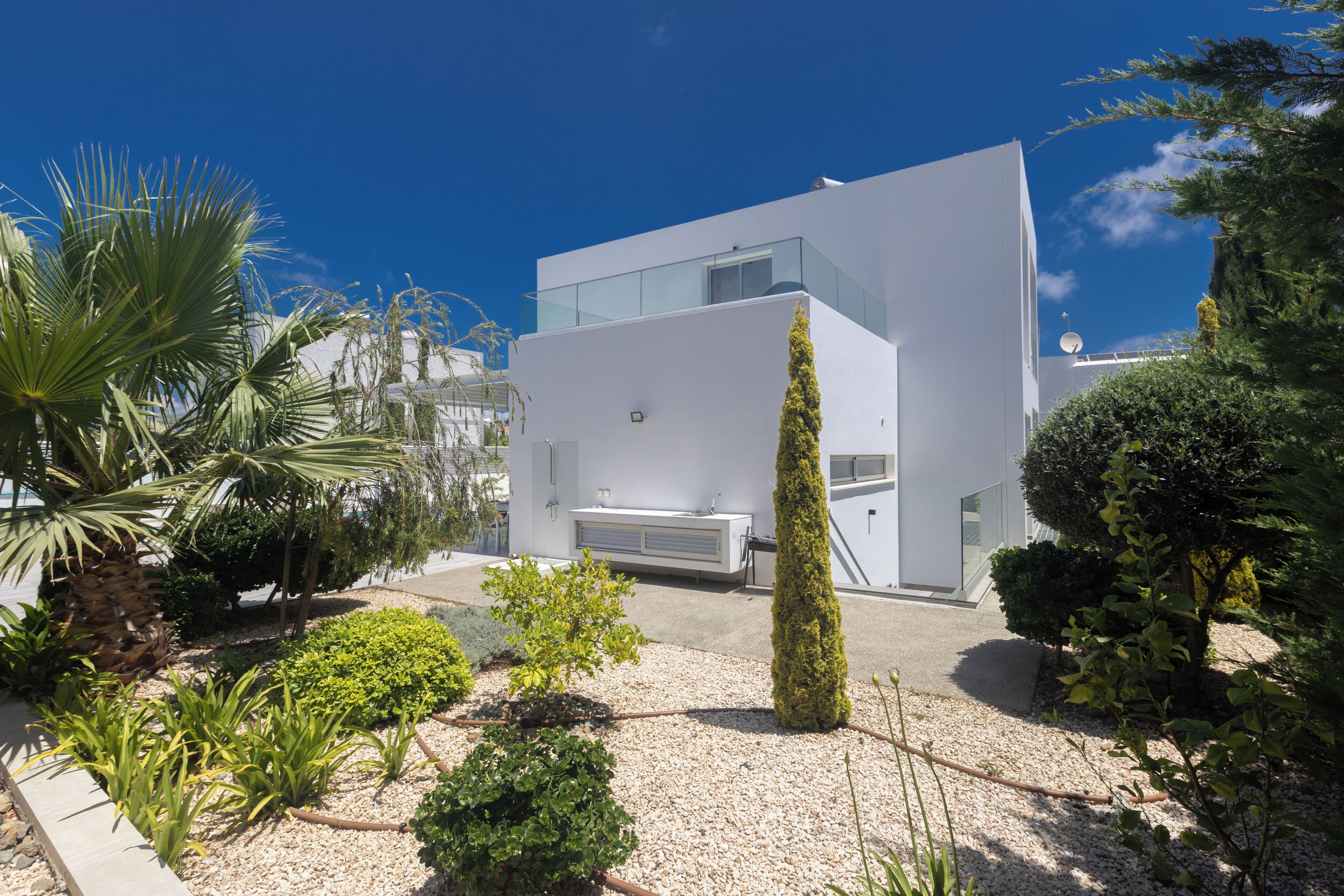 Un edifici modern i blanc amb teulada plana, envoltat d'un jardí de grava amb arbustos diversos i un arbre alt i cònic.