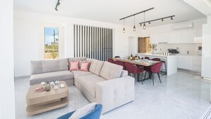 A modern living room with a sectional sofa, a dining area with red chairs, and a kitchen with white cabinets. - 4 Bedroom Villa in Protaras Center - Protaras Vie Bleu Villa Vb3 (Paralimni)