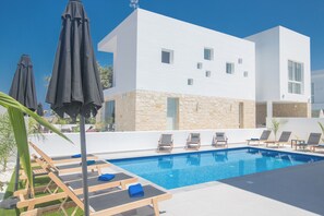A modern pool area with lounge chairs and umbrellas, a stone facade building, and a clear blue sky. - 4 Bedroom Villa in Protaras Center - Protaras Vie Bleu Villa Vb3 (Paralimni)