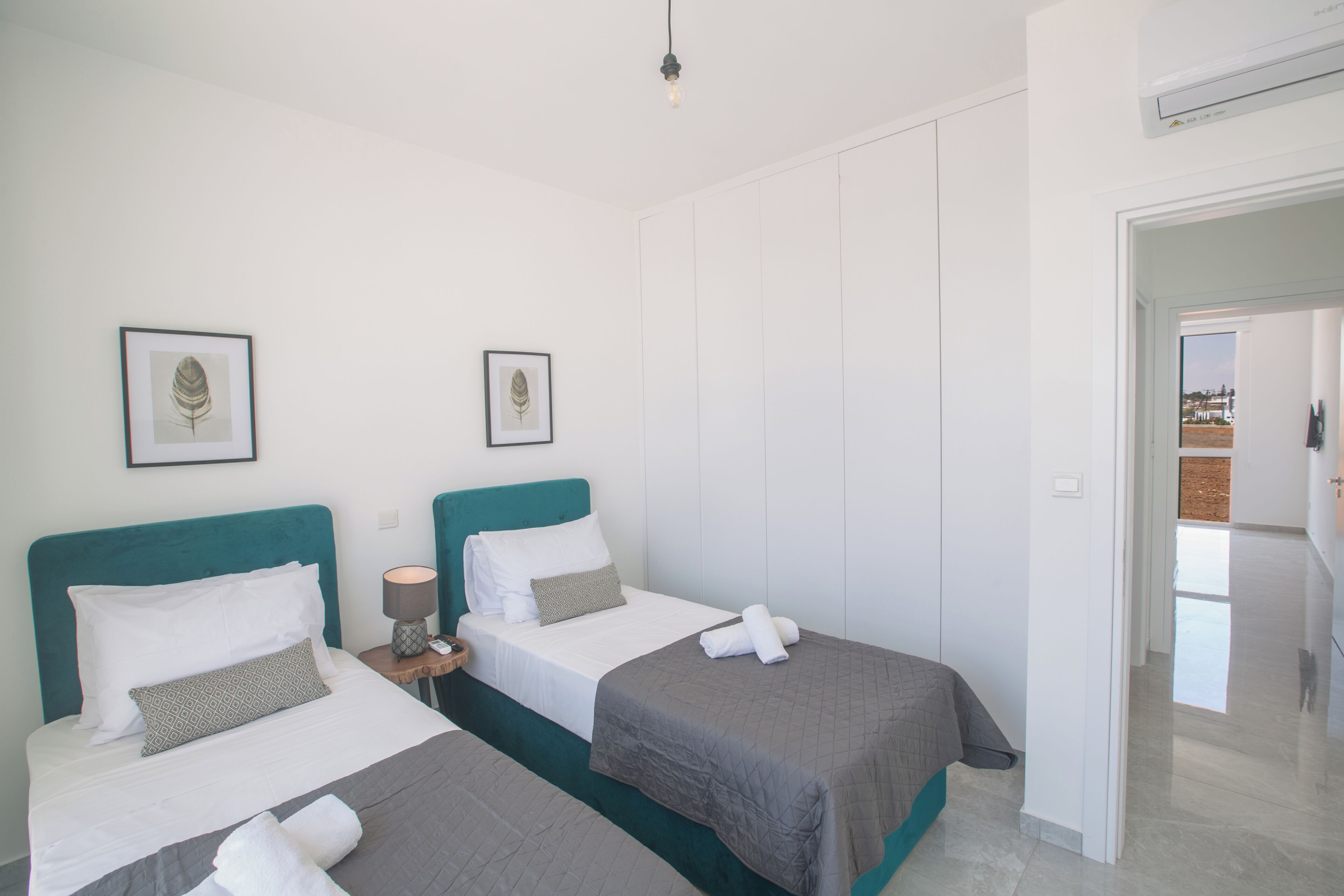 A hotel room with two beds, each with a teal headboard, white bedding, and a gray blanket. There are framed leaf artworks on the wall, a bedside lamp, and a door leading to a balcony.