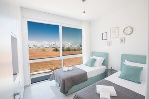 A modern bedroom with two beds, large windows, and a view of a building and landscape. - 4 Bedroom Villa in Protaras Center - Protaras Vie Bleu Villa Vb3 (Paralimni)