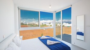 A modern hotel room with a large bed, a view of a cityscape through large windows, and a balcony with a view of the beach. - Protaras Holiday Villa Bp3 (Paralimni)