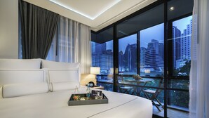 A modern hotel room with a large bed, a bedside table, a lamp, and a view of the cityscape through large windows. - Tastoria Collection Hotel Sukhumvit (Bangkok)
