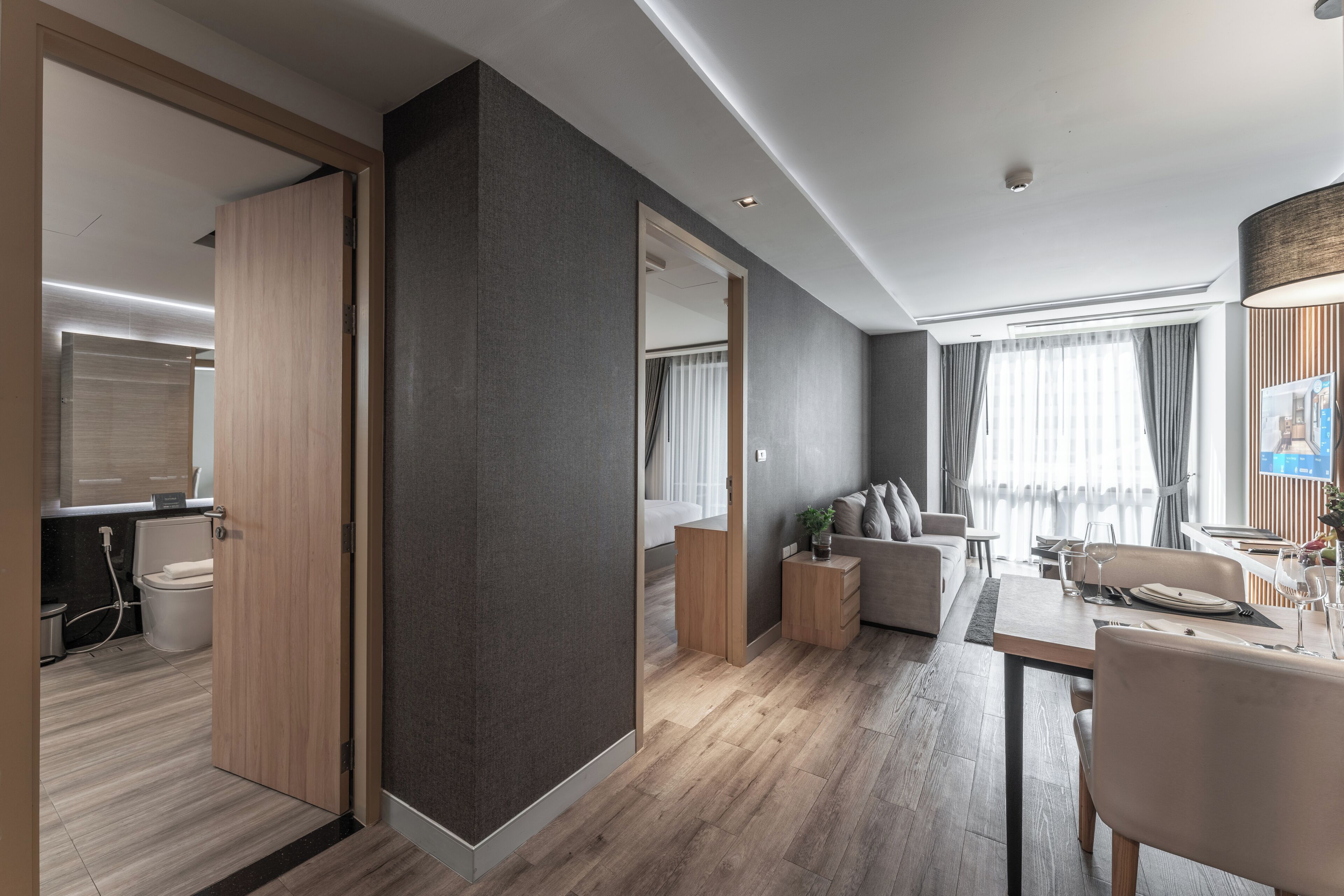 A modern hotel room with a wooden door, a grey accent wall, a dining area with a table and chairs, a sofa, and a view of the city through large windows.