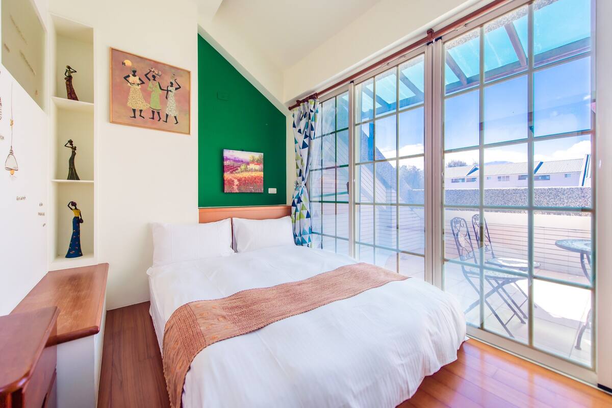 A bedroom with a large bed, a wooden nightstand, a green accent wall, and a view of the outdoors through a sliding glass door.