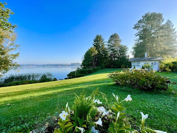 A serene lakeside property with a well-maintained lawn, a small white house, and dense trees.