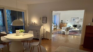 A modern living room with a round dining table, white chairs, a white sofa, and a coffee table. There is a fireplace, a plant, and a framed picture on the wall. - Haus Se[e]renity - Cozy house by the lake (Hungen)