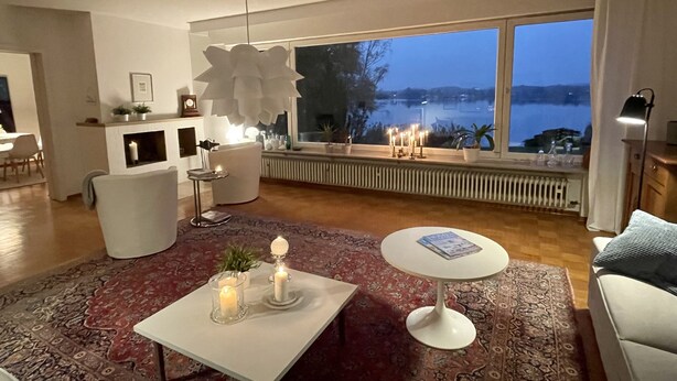 A modern living room with a large window, a white sofa, a coffee table with candles, and a view of a body of water. - Haus Se[e]renity - Cozy house by the lake (Hungen)