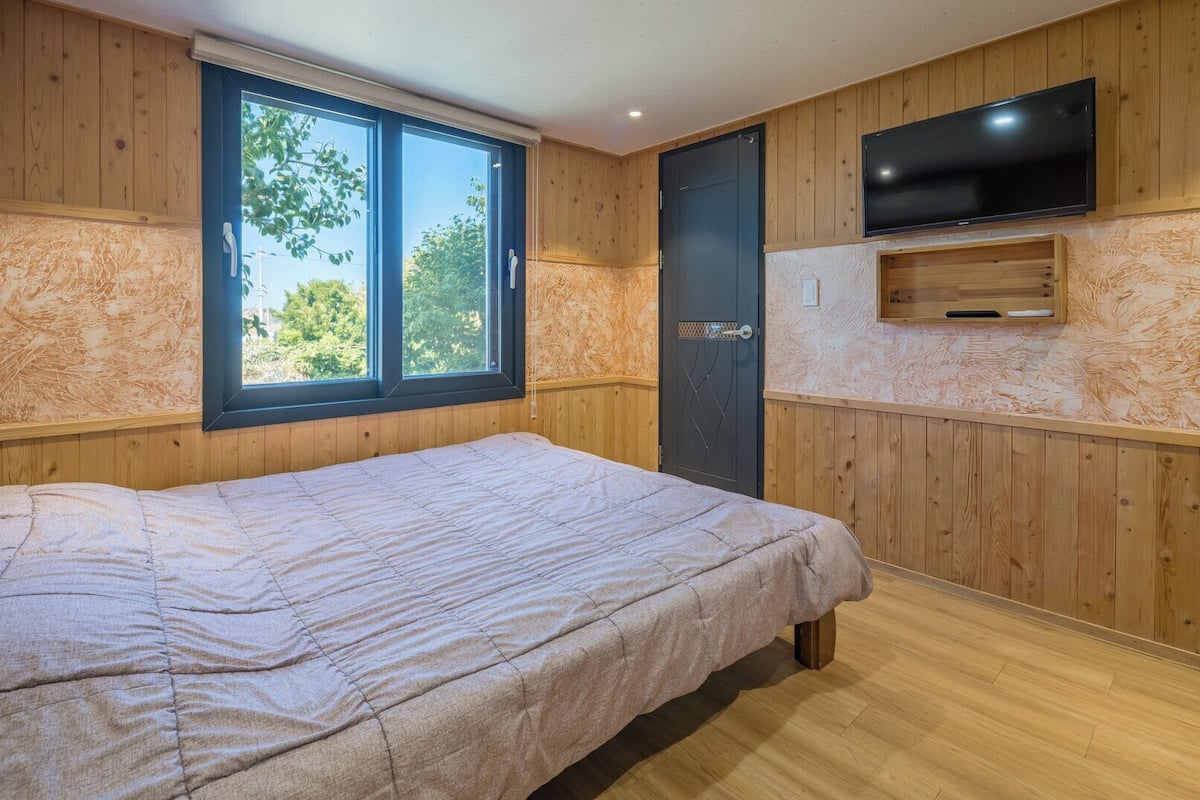 A bedroom with a bed, a window, a door, and a TV mounted on the wall.