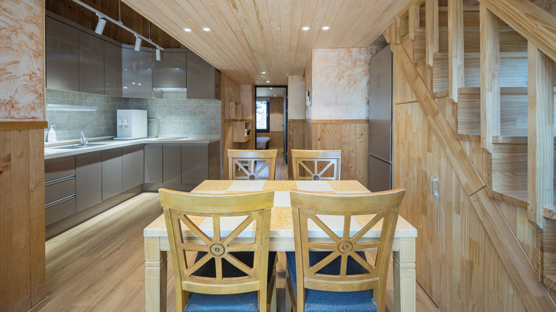 A modern kitchen with wooden cabinetry, a central island, and a dining area with two wooden chairs and a table.