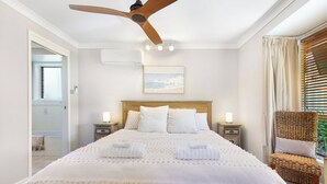 Un dormitori amb llit de fusta, roba de llit blanca, ventilador de sostre, taulells amb làmpades i una cadira de vimet.