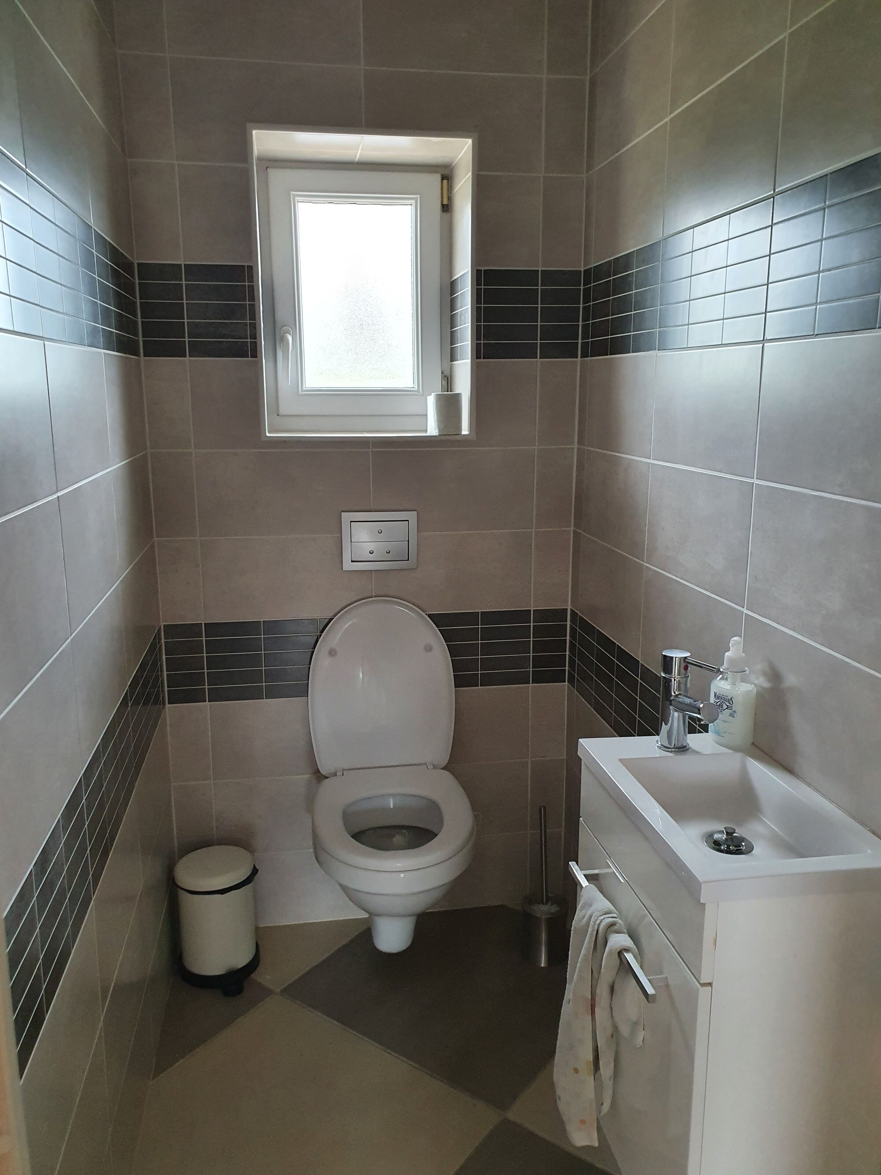 A bathroom with a toilet, a window, a sink, and a trash bin.