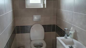 A bathroom with a toilet, a window, a sink, and a trash bin.
