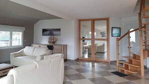 A spacious living room with a large white sofa, a wooden coffee table, and a ship model on a console table. A wooden staircase with a railing leads to a door with glass panels. - Charming, bright, spacious house (sleeps 12) 2 mns from the beach (Locquirec)
