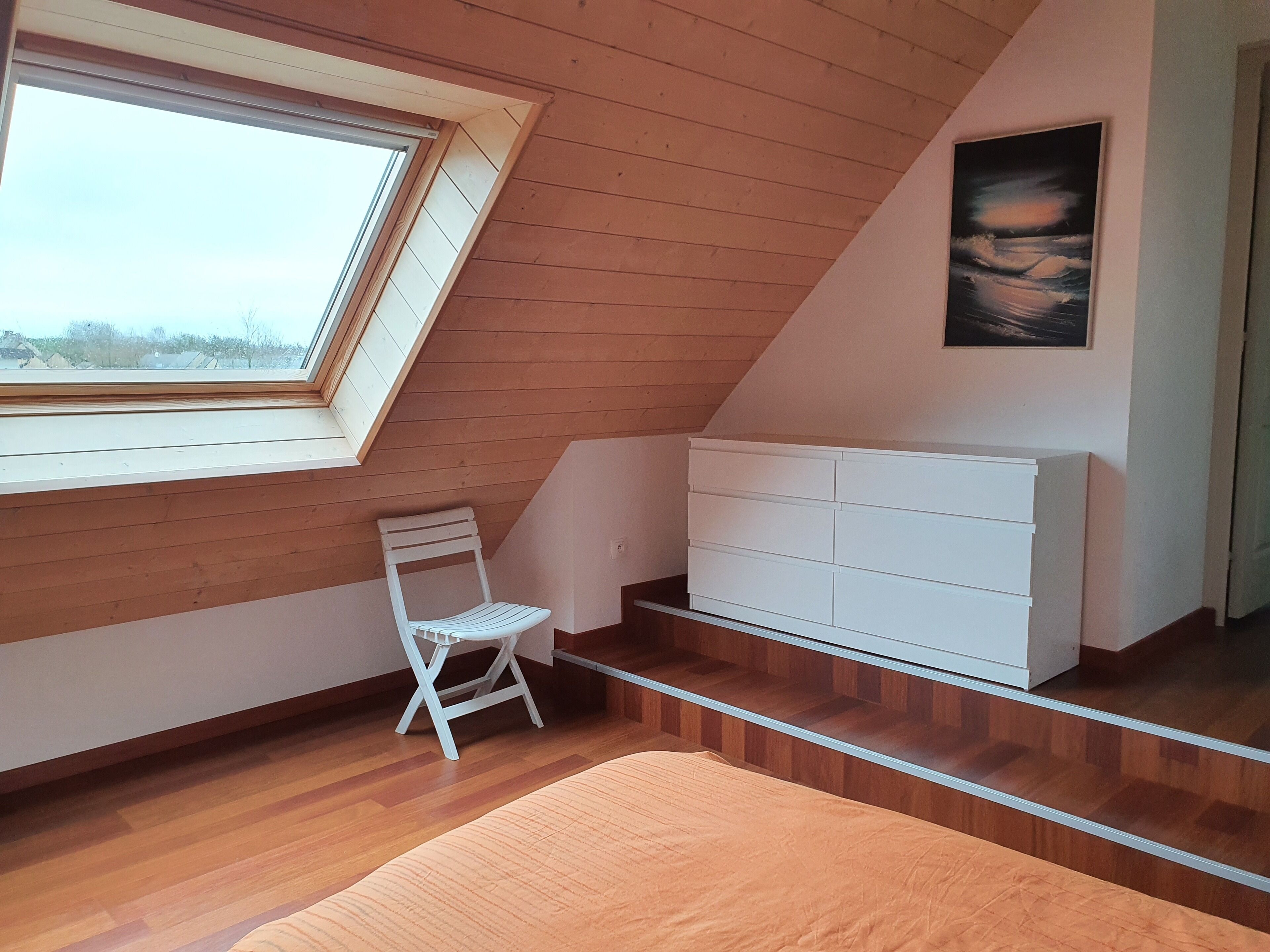 Een kamer met schuine wand, een witte radiator, een witte stoel en een raam met uitzicht op bomen.