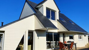 A house with a sloped roof, large windows, and a deck with outdoor furniture. - Charming, bright, spacious house (sleeps 12) 2 mns from the beach (Locquirec)