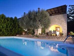 A swimming pool with a stone building in the background, a patio with outdoor furniture, and a tree.
