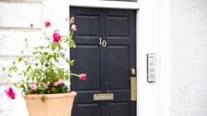 Een zwarte deur met het nummer 10, een bloempot met roze bloemen en een intercom bevestigd aan de muur.