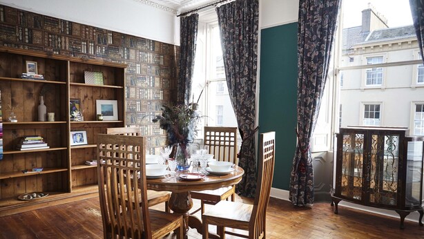 Een eetkamer met een houten tafel gedekt voor vier personen, omringd door houten stoelen. Een boekenkast vol boeken en decoratieve objecten bevindt zich aan de linkerkant.