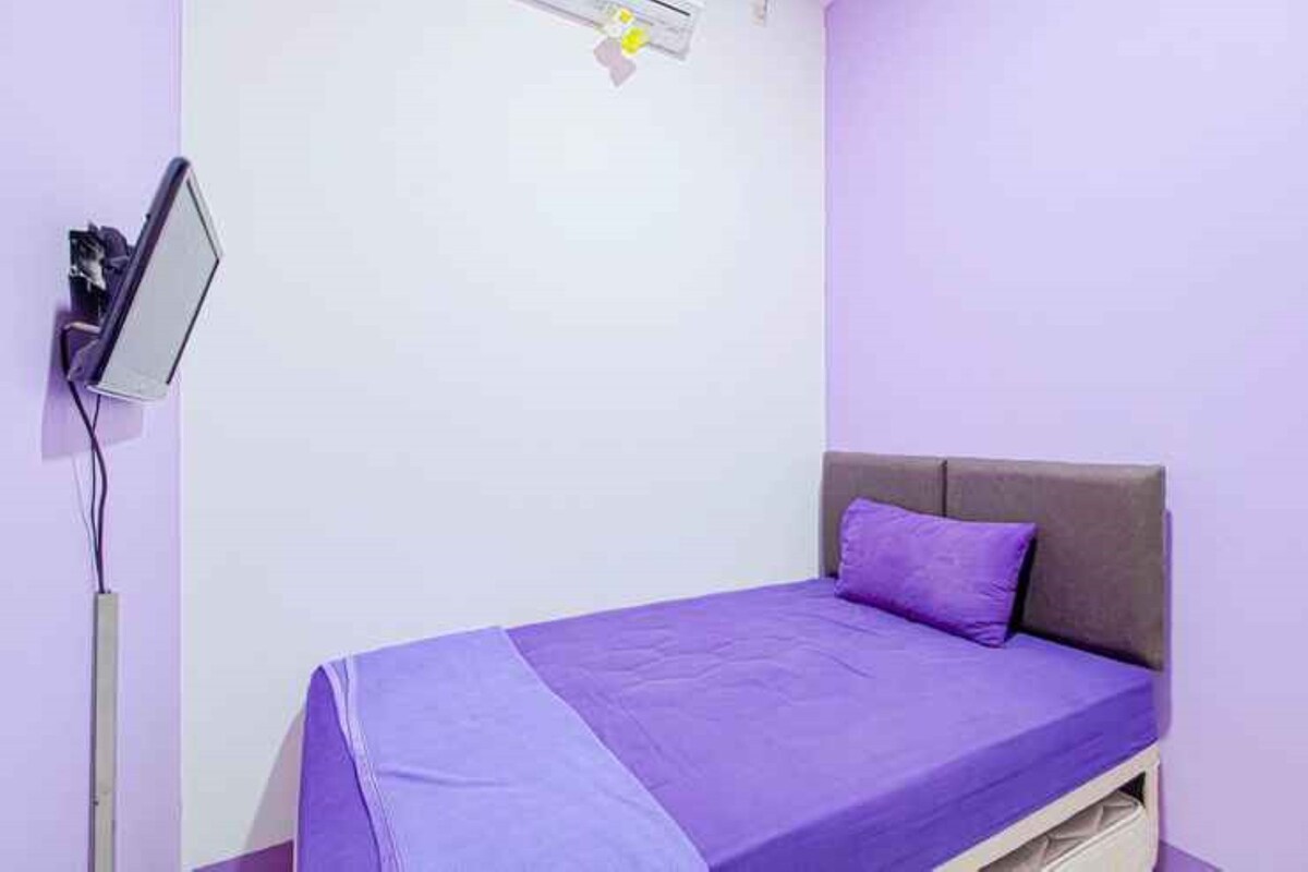 A single bed with a purple bedspread and a headboard, a bedside table, and a wall-mounted TV.