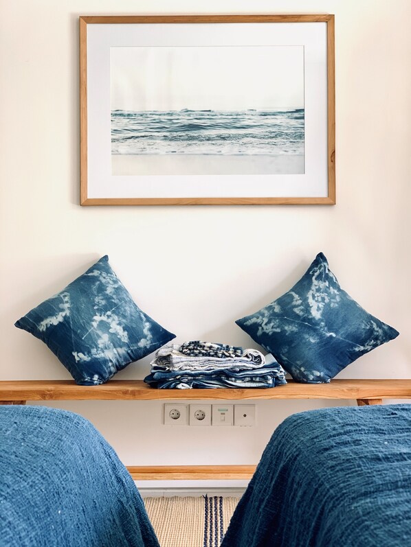 A framed picture of the ocean on a wall, a wooden shelf with folded clothes and two blue pillows, and a bed with blue bedding. - Pelangi 148 Beach Suites (Seminyak)