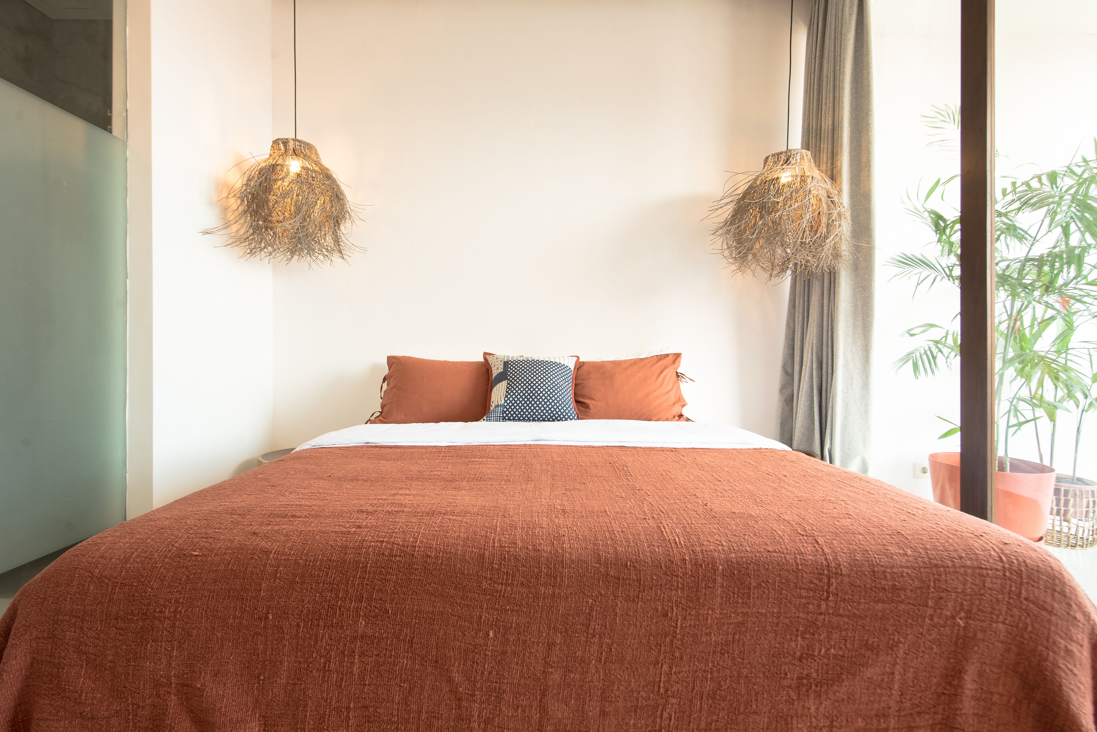 A bedroom with a large bed, two hanging lamps, and a window with a view of greenery.