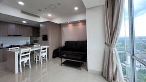 A modern kitchen with white cabinetry, a dining area with a white table and chairs, a black sofa, and a large window with a view of the city. - U Residence 2 by Ana Room (Curug)