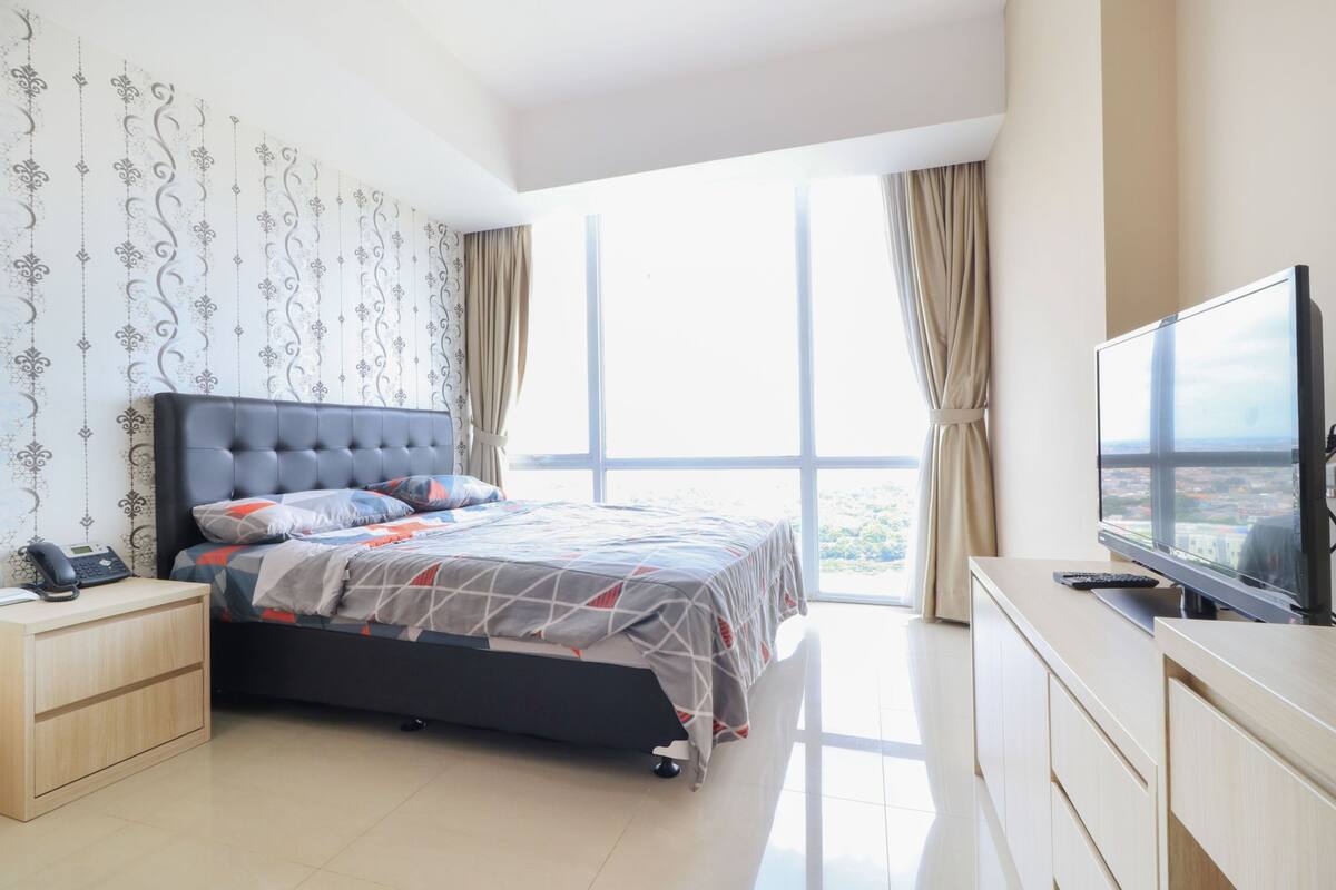 Ein Schlafzimmer mit einem großen Bett, einem Fernseher, einem Nachttisch und Blick auf die Stadt durch große Fenster.