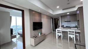 A modern living room with a flat-screen TV, a dining area with white chairs, and a kitchen with wooden cabinets. - U Residence 2 by Ana Room (Curug)