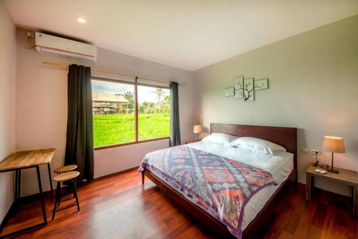 A bedroom with a large bed, a desk, a chair, and a window with a view of a green field.