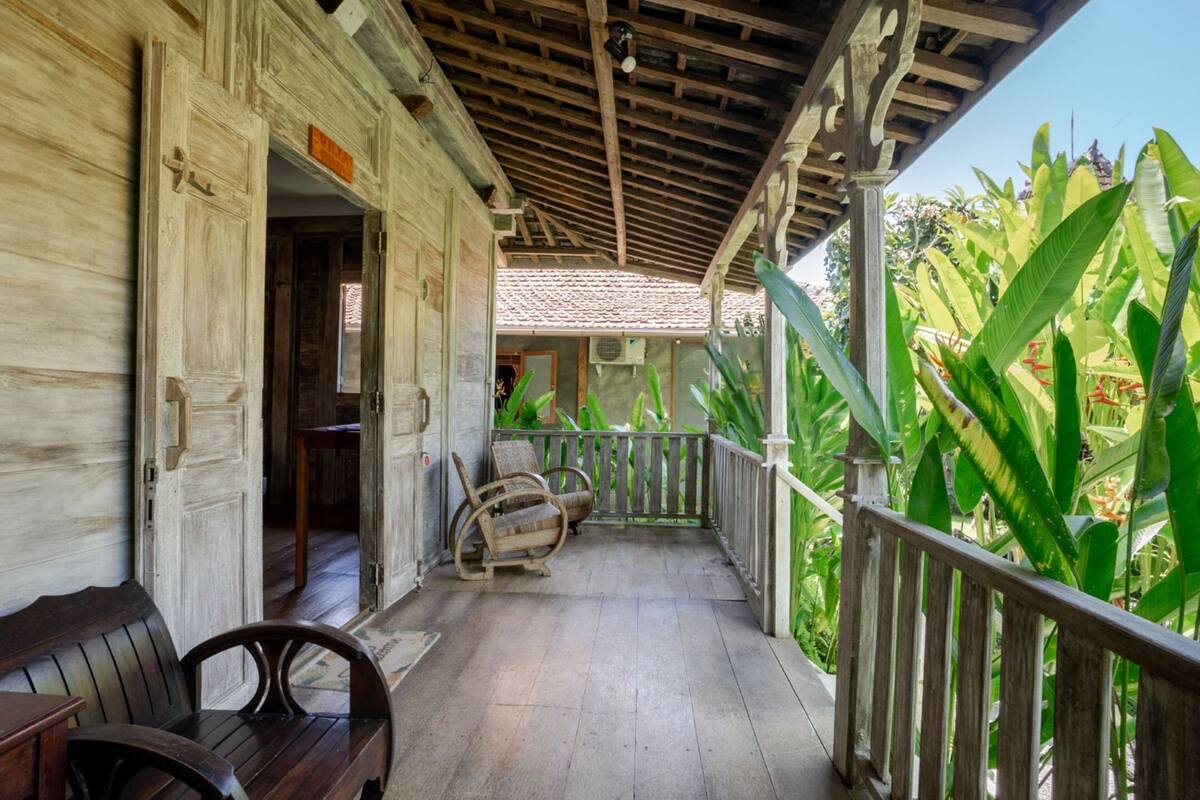A wooden porch with a bench and a rocking chair, surrounded by lush green plants.