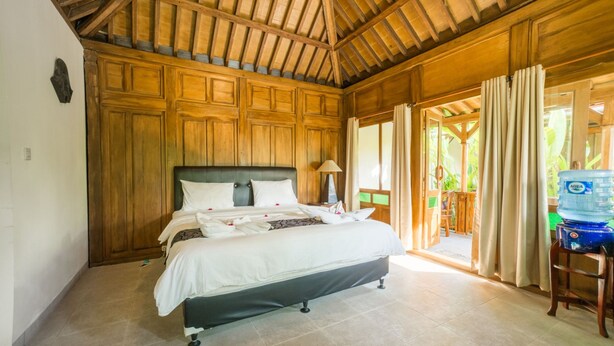 Un dormitorio con techo de madera, una cama con ropa blanca, una mesita de noche con lámpara y vista al exterior por una puerta.
