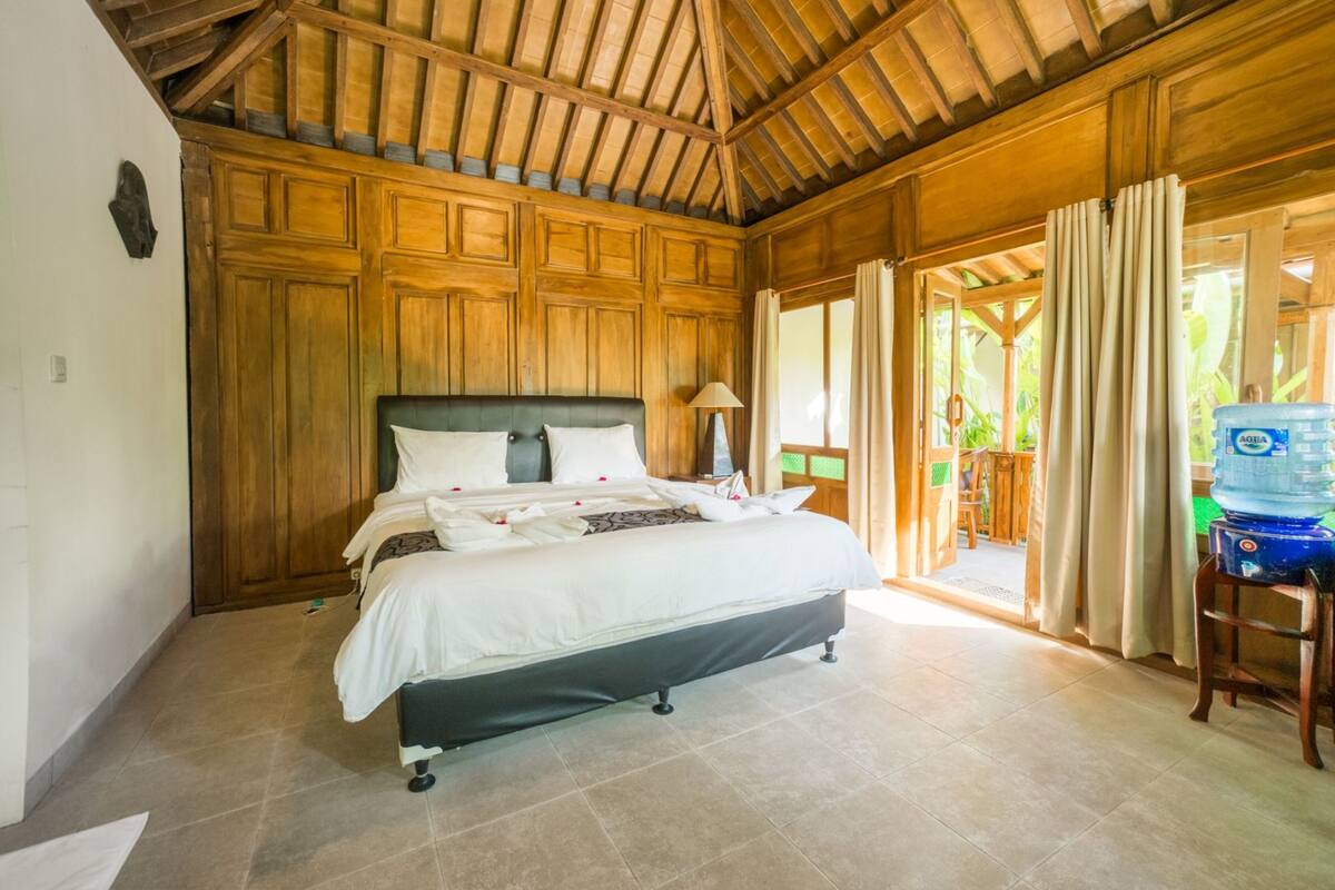A bedroom with a wooden ceiling, a bed with white bedding, a nightstand with a lamp, and a view of the outdoors through a door.