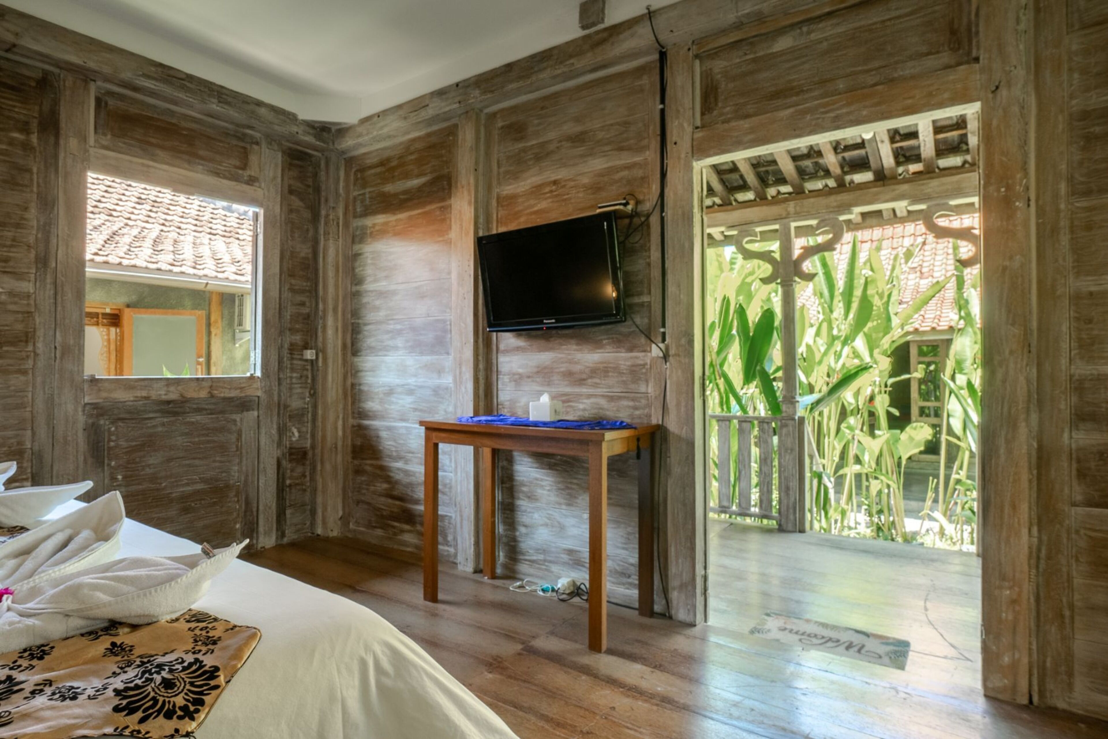 Habitación con paredes de madera, una cama con sábanas blancas, una pequeña mesa con mantel azul, un televisor en la pared y vista a zonas verdes a través de una puerta.