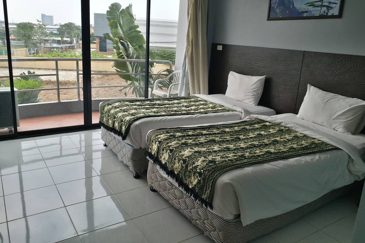 A hotel room with two beds, a large window with a view of buildings and greenery, and a painting on the wall.
