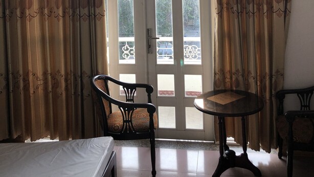 Una habitación con una cama, dos sillas, una mesita y un ventanal grande con cortinas.