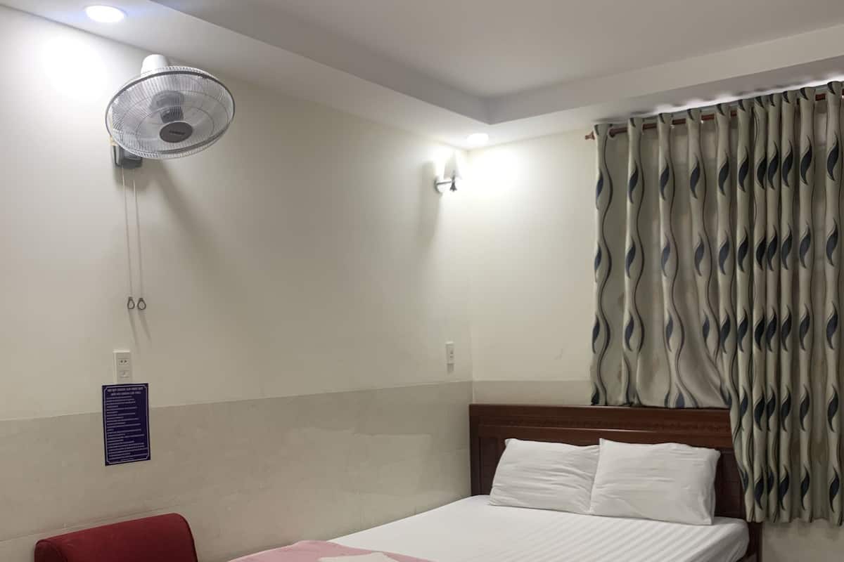 Une chambre d’hôtel avec un lit, une chaise rouge, un ventilateur sur pied et un rideau.