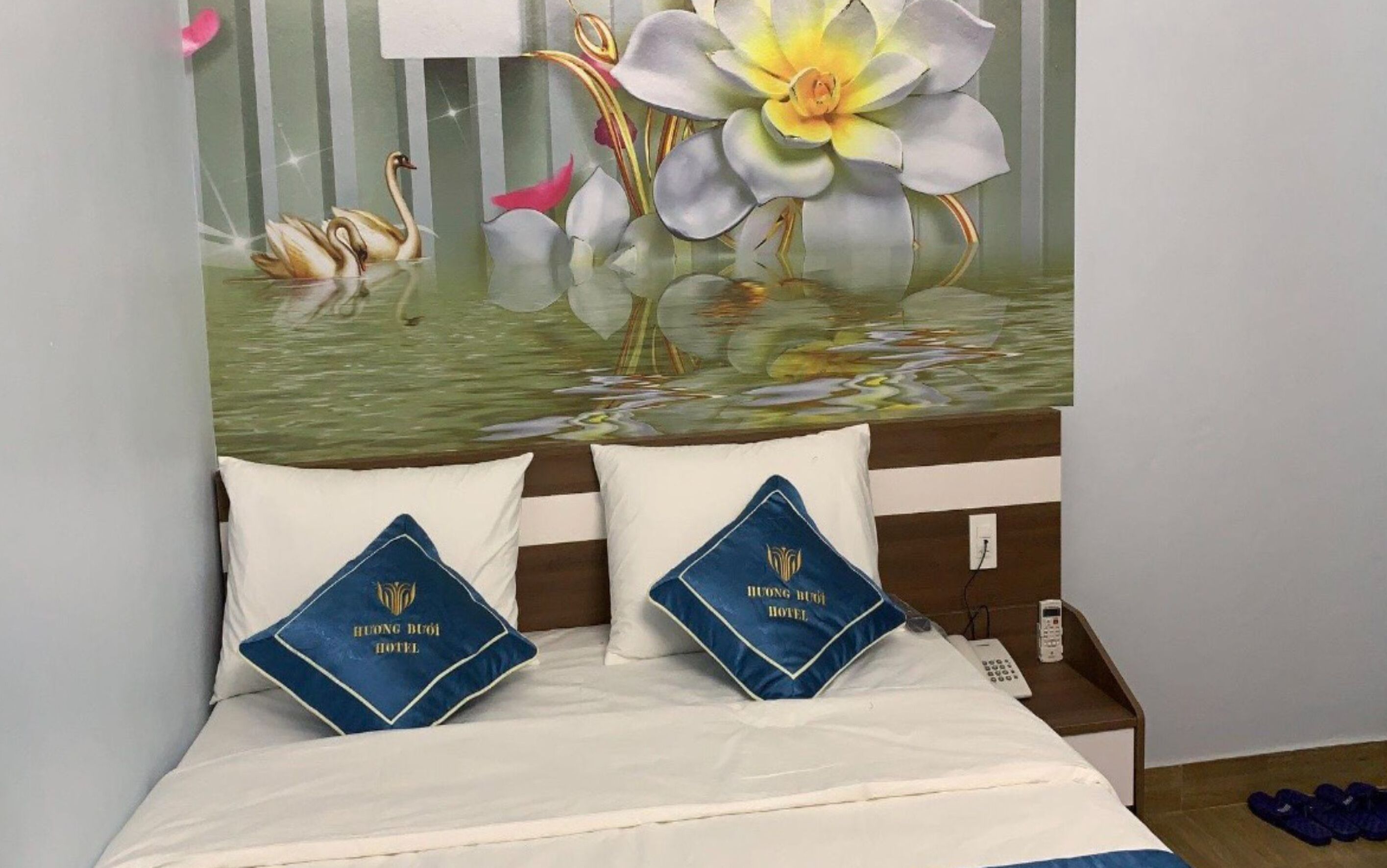 Quarto de hotel com uma cama, duas almofadas com o logotipo do hotel, uma mesa de cabeceira com telefone e um mural de cisnes e flores na parede.