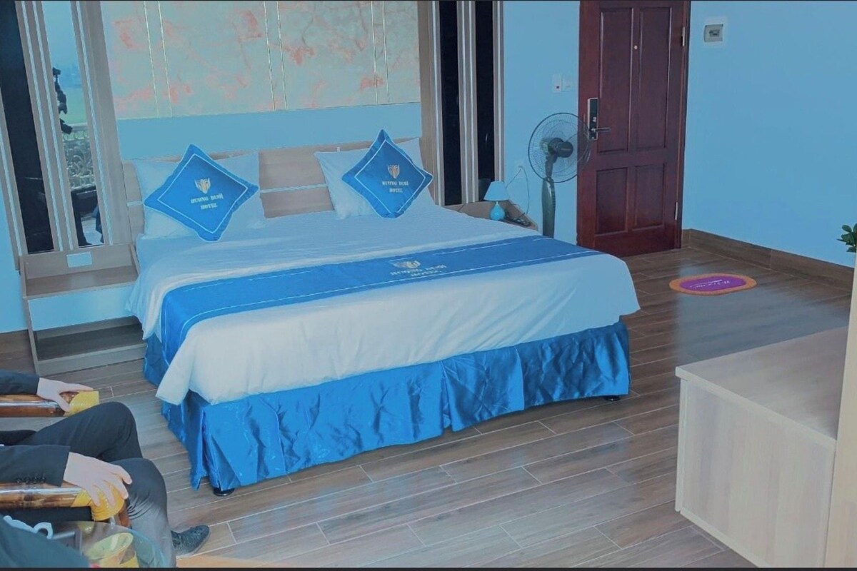 A hotel room with a bed, a desk, a chair, a standing fan, and a door.