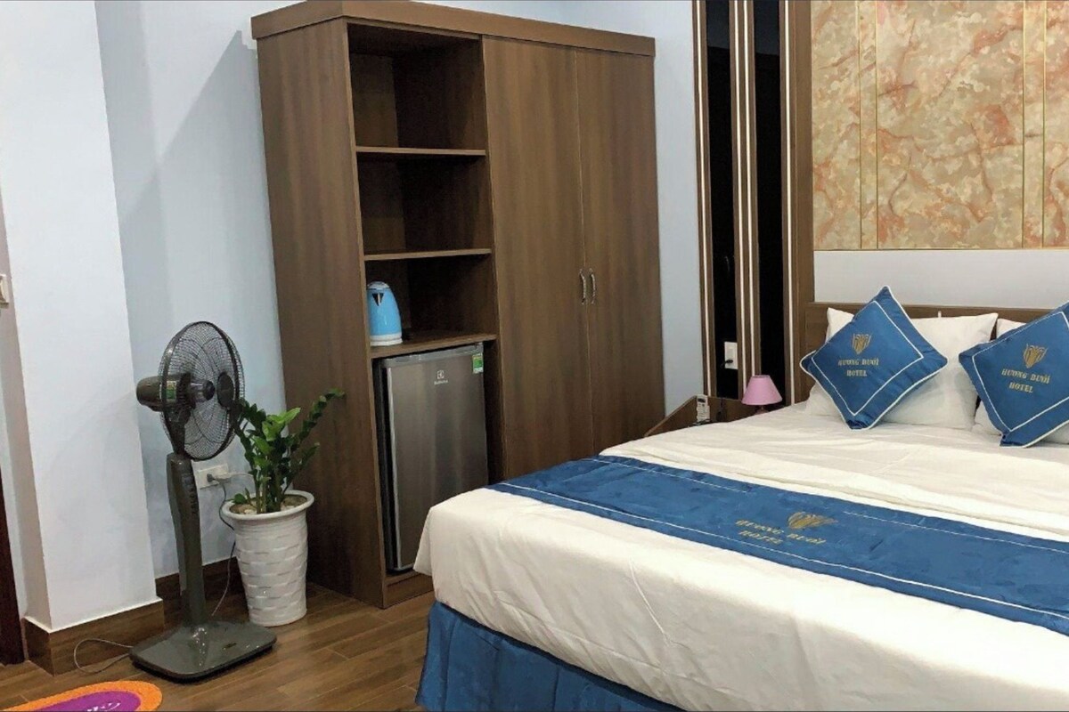 A hotel room with a bed, a wardrobe, a small fridge, a fan, and a potted plant.